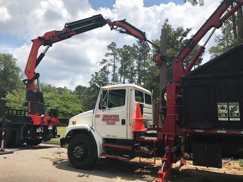 Specialized crane rental truck with dual hydraulic arms positioned for emergency tree removal and debris hauling operations in wooded terrain.