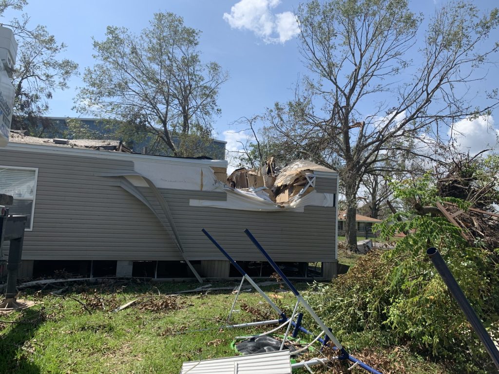 Storm-damaged mobile home with roof and wall destruction revealing interior framing, surrounded by debris from severe weather event.